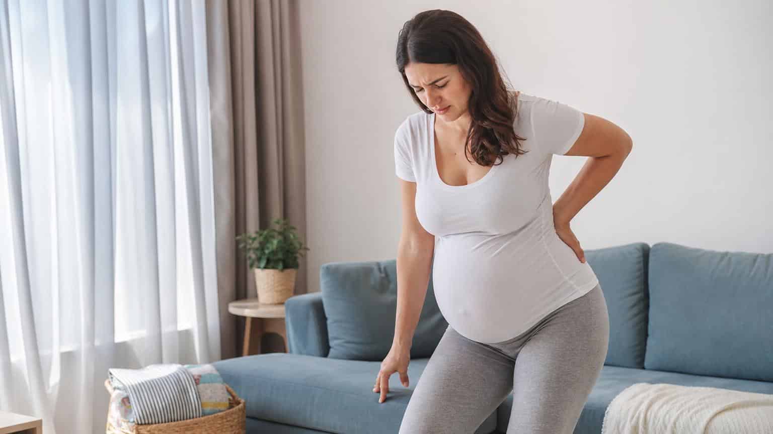 Pregnant woman in a white t-shirt standing in a living room, grimacing while holding her lower back in pain.