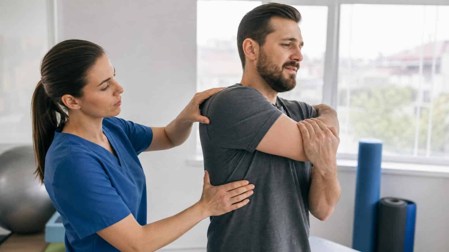 Female physical therapist in blue scrubs assisting a man with a cross-body shoulder stretch in a bright rehabilitation clinic.