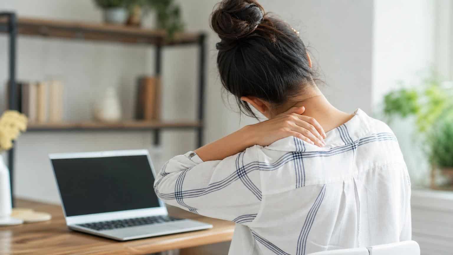 Woman sitting at a desk with a laptop, seen from behind, rubbing her neck and shoulder in discomfort.