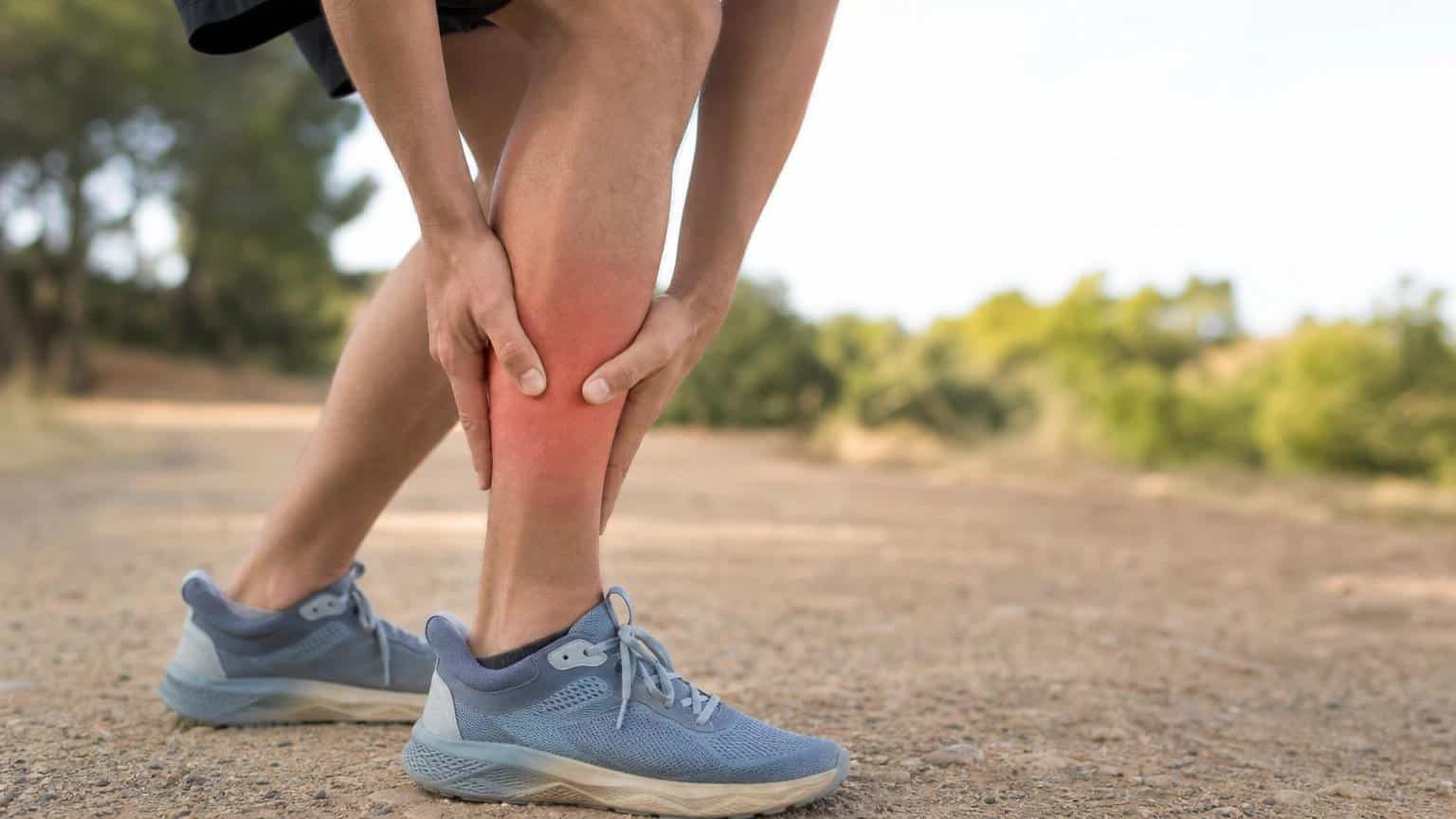 Runner clutching a painful, red-highlighted shin while standing on a dirt path outdoors.