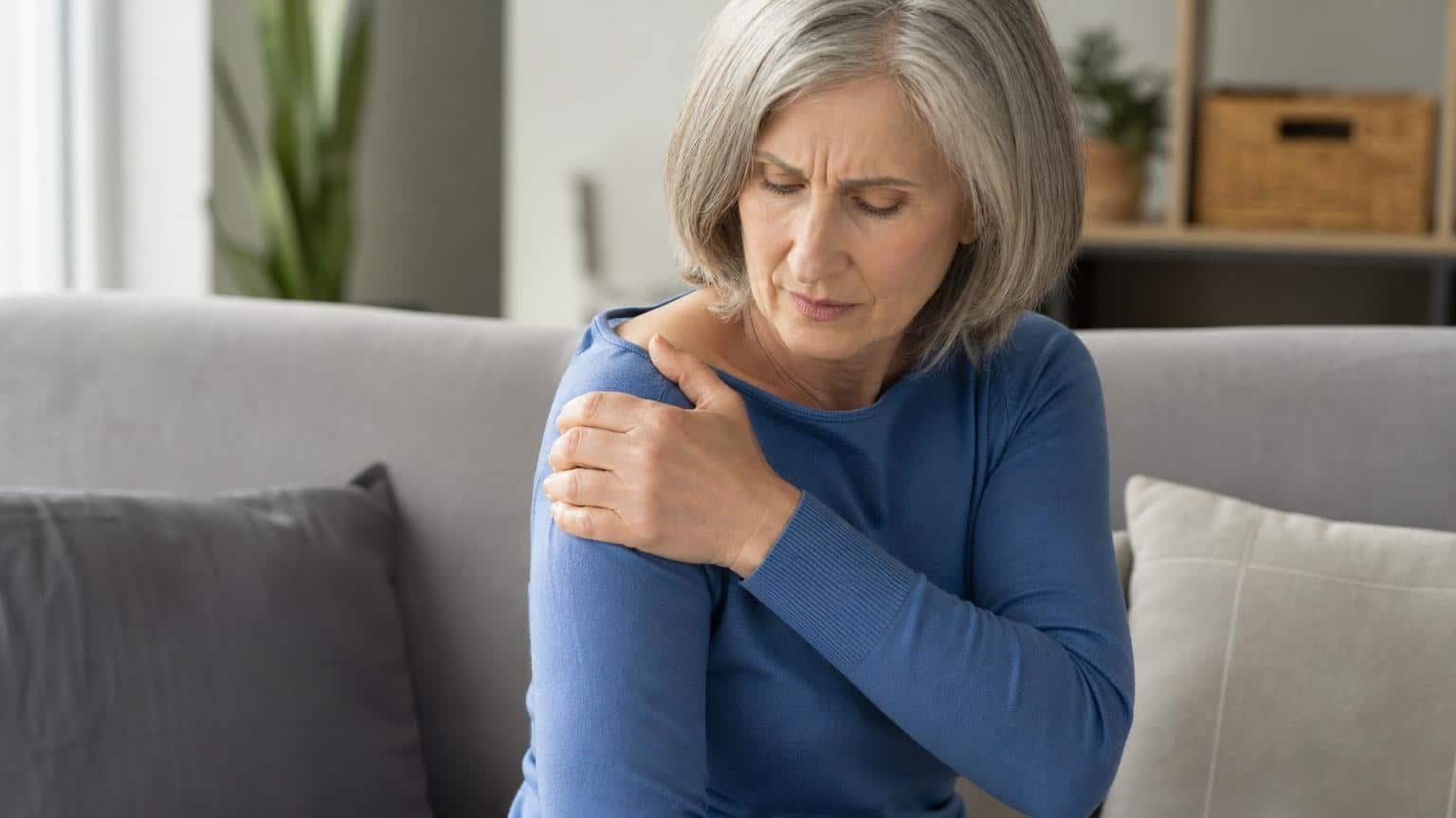Senior woman with grey hair sitting on a sofa, grimacing in pain while holding her right shoulder.